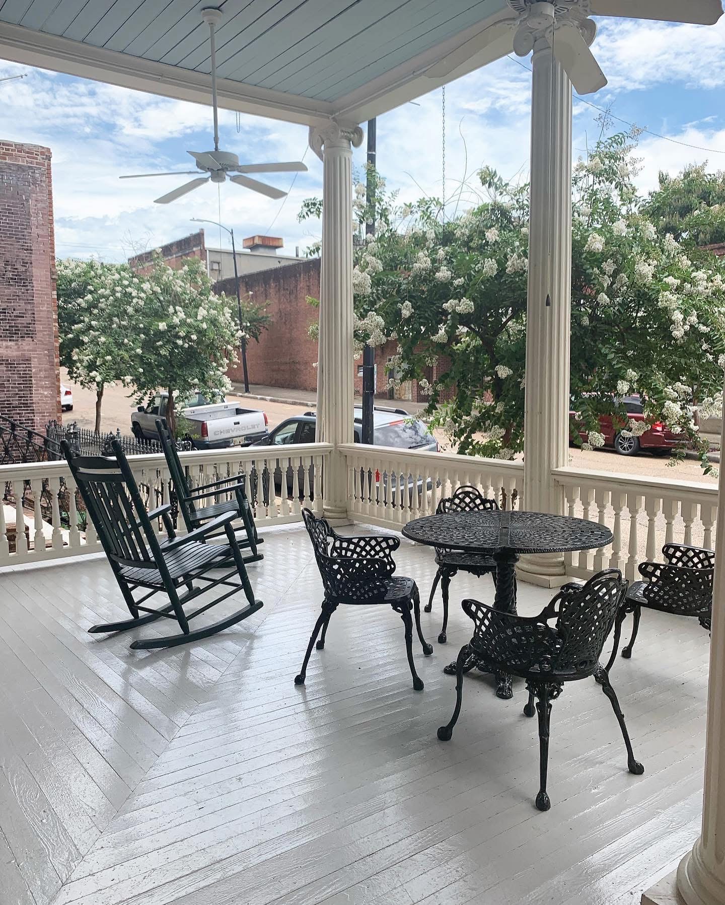 Two rocking chairs and a cast-iron table set on the upper gallery of The Guest House, crepe myrtles in bloom beyond the rail.